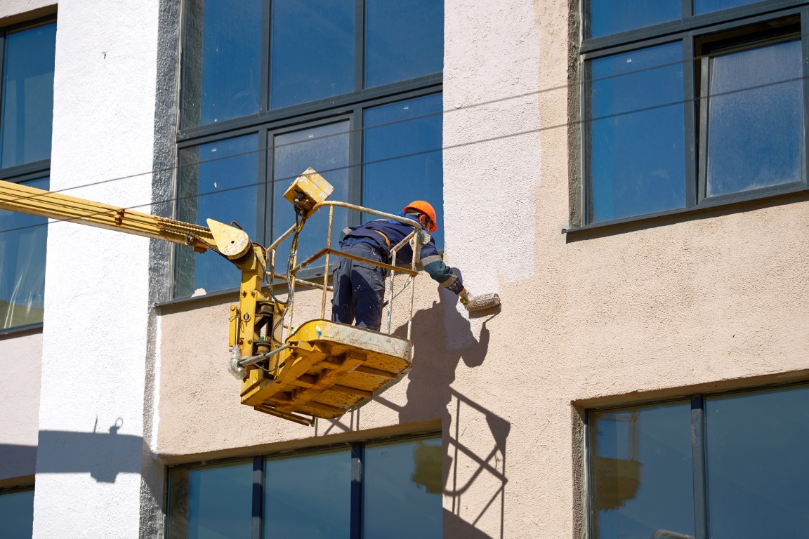 Man paint wall with brush roller in lifting bucket. Painter working on crane platform, paint building wall. Men at work in hardhat paint facade of building at height in lifting cradle. Renewal work.